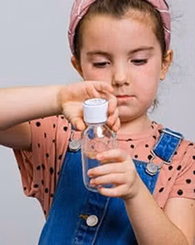 Young girl in denim overalls opening a small clear bottle with a white cap.