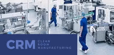 Workers in blue scrubs walk among automated machinery in a clean room manufacturing facility.