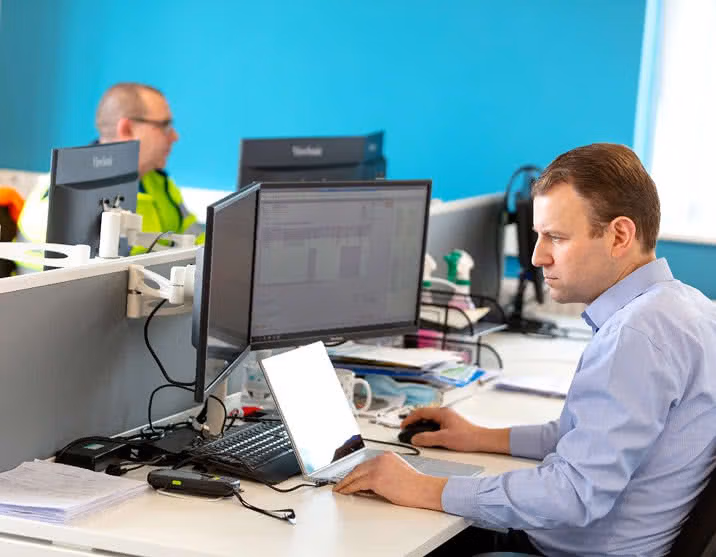 Man in a blue shirt working at a desk with dual monitors, laptop and phone in an office.